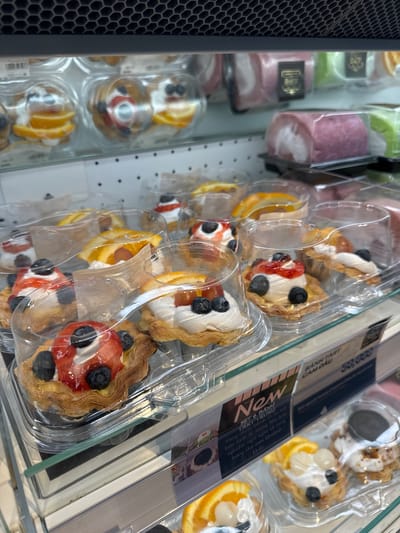The image shows a close-up, eye-level view of pastries displayed in clear plastic containers on shelves inside a commercial establishment, likely a bakery or convenience store. The pastries are varied, including tarts topped with fruit such as strawberries, blueberries, and orange slices, as well as cream and syrup. Some pastries are circular, while others appear to be part of a roll cake. The background displays more packaged pastries, some with colorful frosting and designs, including pink and green roll cakes. A price tag and signage in Vietnamese are visible, with text including "BANH TART CAM DAU" and "NEW," suggesting these are fresh offerings. The lighting is bright, characteristic of retail display cases. The environment appears clean and organized, with a focus on presenting the food items attractively. The specific location of Phu Thuy, Vietnam, is not explicitly indicated in the visual elements of the image itself.