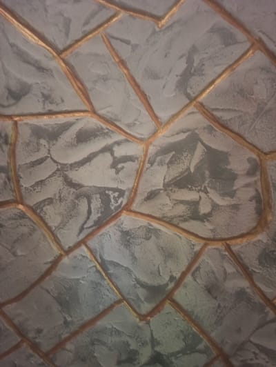 The image shows a close-up of a decorative ceiling or wall that mimics a stone pattern. The "stones" are rendered in shades of grey with a rough, textured surface, and are outlined by raised, golden-brown lines that create a mosaic or cracked earth effect. The lighting appears to be dim, casting subtle shadows that accentuate the texture of the surfaces. The image does not provide any information or context to determine its location in the city of Moldoveanca, Moldova.