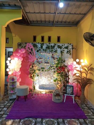 This media file captures a decorated area, likely for a celebration or event, in what appears to be an indoor or semi-outdoor residential setting in Lallatang, Indonesia. The scene is vibrant and festive, with a prominent display featuring balloons, flowers, and a decorative backdrop.

The central focus is a white, ornate baby carriage adorned with a butterfly motif. In front of the carriage, a plush teddy bear sits on a purple rug. A framed photograph of a young child in a pink dress is displayed on a white easel to the right of the carriage. A small, circular, grey stool is positioned to the left of the carriage.

The backdrop consists of a sequined, shimmering material layered with a white fabric and decorated with artificial green vines. A large pink butterfly cutout is attached to the left side of the backdrop. A balloon arch, composed of pink and white balloons, curves from the left side of the frame towards the backdrop. Another cluster of pink and white balloons is situated on the right side of the scene.

Decorative floral arrangements, featuring pink, white, and red flowers, are placed on either side of the baby carriage, adding to the festive atmosphere. Two floor lamps with multiple white globe lights are also visible, one on the left and another on the right, contributing to the illumination of the scene. A tall, brown vase with dried, feathery foliage stands to the right.

The walls are painted a bright yellow, and the ceiling is constructed of corrugated metal. The floor is tiled with a patterned design. The overall impression is one of a happy and celebratory occasion, possibly a baby shower, naming ceremony, or birthday party.

Visible text on the backdrop reads "TASYAKURAN AQIQAH" and "Kiara Elmadina Maynarani". The time of day appears to be evening or night, indicated by the artificial lighting.