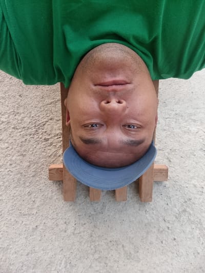 The image is an upside-down, close-up selfie of a man wearing a green t-shirt and a light blue baseball cap. He is lying on what appears to be a wooden structure, possibly a bench, which is resting on a rough, textured surface, likely concrete or cobblestone. The background is predominantly this rough texture. The lighting suggests it is daytime, possibly outdoors. The man's face is the central focus, with his eyes visible and looking towards the camera. His expression is neutral. The image was taken in Tash-Dobo, Kyrgyzstan.