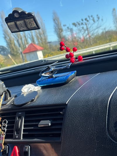 Inside a car, various items are attached to the dashboard and windshield. Red berries hang near a blue device clipped onto the dashboard. A chrome clip holds something next to it. Below, a circular object with a tissue attached is visible. On the windshield, a decorative, rectangular item with symbols is affixed. The car's air vents and controls are partially visible, along with a decorative keychain displaying Audi's logo.