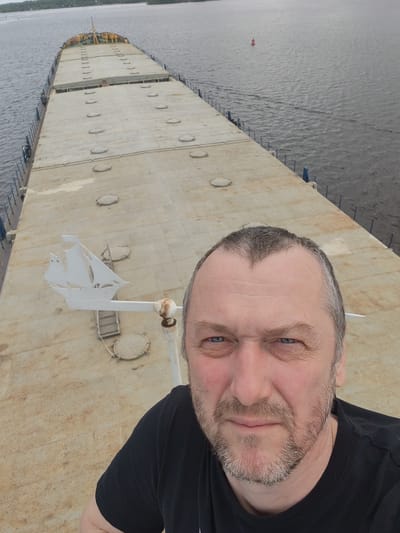 A man with fair skin, graying hair, and a beard stands on the deck of a large, flat-topped barge or ship. He wears a black t-shirt and is looking directly at the camera. Behind him is a long, rectangular expanse of the barge deck, marked with a series of circular access hatches. A white, decorative ship-shaped weather vane is visible on the left side of the barge, near the man. The barge is positioned on a body of water, with a shoreline visible in the distance. A red navigational buoy is visible in the water. Novodevich'e, Russia is implied by the scene.