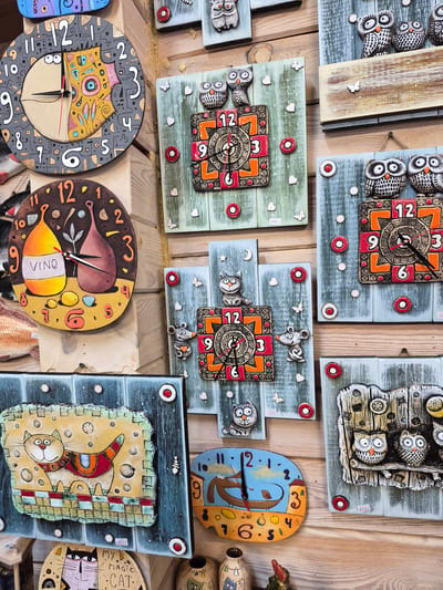 The image depicts a collection of handmade ceramic clocks displayed on a wooden wall, likely in a shop or market stall in Krasny Klyuch, Russia. The clocks are varied in design, featuring whimsical figures such as owls, cats, and mice, as well as more abstract patterns. Some clocks have a traditional round face with Arabic numerals, while others have square or irregularly shaped designs.
Prominent among the clocks are several with owl motifs. One large round clock, in the upper left, is gray with black numbers and a colorful, abstract face. Below it, a circular clock with a brown background features two wine bottles and the word "VINO". To the right, a series of square or rectangular clocks are arranged vertically. These include clocks with owls, hearts, and decorative patterns in oranges and reds. One clock features a charming cat in a colorful scarf, and another shows a blue background with a boat and a bird. A clock with a mouse figure is also visible.
The wooden wall serves as a rustic backdrop, with visible grain and texture. The lighting appears to be natural or from overhead, casting a soft illumination on the objects. There are no people visible in the image. The scene is static, presenting a still life of decorative items. Small price tags are visible on some of the clocks.