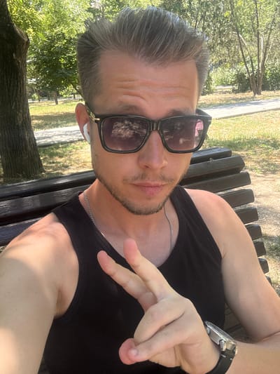 A man is sitting on a park bench, taking a selfie. He has short, graying hair, is wearing sunglasses and a black tank top, and has a silver chain around his neck. He is also wearing a watch on his left wrist and has a small beard. He is making a peace sign with his right hand. The background is a park with trees and sunlight filtering through the leaves. There is a paved path in the background. The man is positioned in the foreground, taking up most of the frame. The overall scene is casual and relaxed, suggesting a pleasant day outdoors.