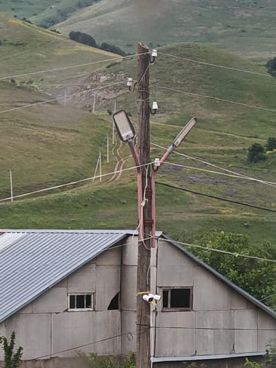 This image depicts a rural scene in Bazum, Armenia, with a focus on a utility pole and a nearby building. The utility pole, made of wood, stands prominently in the foreground, supporting two streetlights and several electrical wires. Ceramic insulators are visible on the pole, securing the wires. A security camera is mounted on the pole, facing towards the building.

The building, constructed with large, textured concrete blocks, has a corrugated metal roof and two windows. One window on the left is small and appears to be a standard residential window, while the window on the right is larger and darker, suggesting it might be an opening or a window with no glass. A portion of the building's wall to the right of the right window appears damaged or incomplete.

Behind the utility pole and building, the landscape consists of rolling, grassy hills. A dirt road winds its way up one of the hills, with several smaller poles and wires marking its path. The vegetation on the hills is predominantly green grass, with some patches of darker green foliage and small purple flowers visible in the mid-ground. The sky is not visible in the frame.

The lighting suggests it is daytime, likely overcast or late afternoon, as there are no strong shadows. The overall atmosphere is one of a remote, possibly underdeveloped, rural area. There is no text visible in the image. No people are present. The scene appears to be static, with no specific activity taking place.