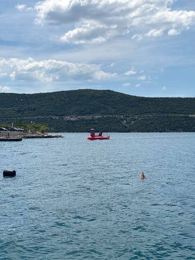 The image shows a vertical landscape view. On the left, there is a cloudy sky. Next to it, there is a green mountain range. The right part of the image depicts a body of water, likely the Adriatic Sea, in Neum, Bosnia and Herzegovina. In the water, there is a red boat with two people in it and an orange object floating nearby. Near the bottom of the image, there is a black floating object. The coastline, with some vehicles and structures, is visible between the mountain range and the sea.