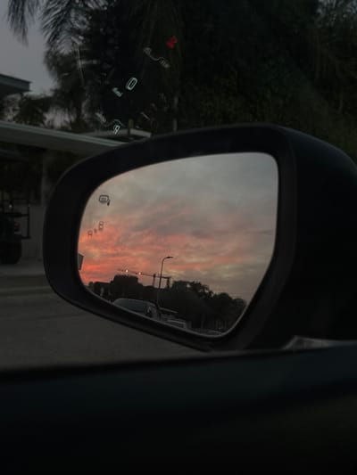 The image depicts an exterior view from the interior of a vehicle, facing the driver's side rearview mirror, within the Emek Izrael Regional Council, Israel, during either dusk or dawn. The prominent feature is the vehicle's side mirror, which reflects a vibrant sunset sky exhibiting strong orange, pink, and grey cloud formations. Within the mirror's reflection, a street scene is visible, including a partial view of a dark pick-up truck, multiple streetlights, a large construction crane silhouetted against the sky, and various trees and building structures. Above the mirror, through the vehicle's windshield, faint heads-up display (HUD) projections are discernible, including numerical readouts (one clearly showing "0") and a red warning indicator. Outside the vehicle, beyond the windshield, dark palm fronds and other dense green foliage are visible. To the left, a portion of a white building with an attached carport or shaded area is present, along with the rear section of another dark vehicle. The scene is devoid of any individuals.