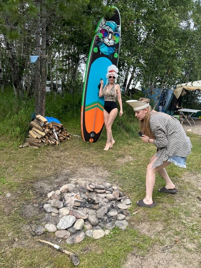 The image depicts an outdoor scene with two individuals in a grassy area, likely a campsite, near a body of water. The location is identified as Tolbazikha, Russia.
In the foreground, a campfire pit is visible, surrounded by stones and ash. A small pile of chopped firewood is stacked to the left of the pit, with a tool resembling a saw leaning against it.
One person, identified as a woman, stands to the left of the frame, leaning against a large, colorful stand-up paddleboard. She is wearing a leopard-print swimsuit, a white wig, sunglasses, and light-colored shoes.
The second person, also a woman, is positioned in the middle ground to the right, facing away from the viewer. She is wearing a plaid jacket over a denim skirt and dark slides. She is looking down at a mobile phone in her hands.
In the background, trees and vegetation are abundant, with a glimpse of a tent and a portable table set up in a camping area. A blue garment is hanging from a clothesline strung between two trees. The overall environment suggests a natural, recreational setting.