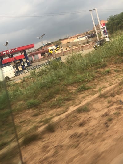 A gas station is visible with red pumps and a white truck. The station appears to be located near a road or path made of dirt or sand, and grassy vegetation borders the road. Further back, there's a row of buildings, possibly a shopping complex, and the sky is overcast. The perspective suggests the photo was taken from a moving vehicle.