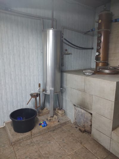 This media file depicts an interior scene, likely in a workshop or utility area, with an industrial distillation apparatus as the central subject. The apparatus includes a copper cylindrical boiler or column, with gauges and valves visible, connected to a large, horizontal stainless steel tank. Copper piping and black hoses are integrated into the system.
The surrounding environment features textured walls, possibly wooden panels on the left and tiled walls and floor on the right. A section of the tiled wall appears to be an opening or recess, revealing a rougher, possibly brick or stone interior.
On the floor to the right of the apparatus, a black plastic tub is present, alongside a small pile of colorful plastic bottle caps.
The lighting suggests an indoor setting, with no clear indication of time of day or weather conditions. The overall impression is one of a functional, perhaps slightly cluttered, operational space. No people are visible in the media file. No discernible text is present. The location context provided is Kochmar, Bulgaria.