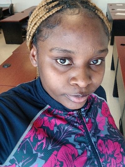 A close-up portrait of a person with dark skin and braided hair. The braids are blonde at the ends and darken toward the scalp. The person's eyes are brown, and they are looking directly at the camera. They are wearing a dark jacket with a bright pink and gray floral pattern. The background is slightly blurred, showing office furniture with dark brown tones.