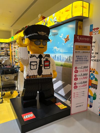 A large, brick-built LEGO minifigure, designed as a pilot, stands prominently in a well-lit retail environment. The pilot figure wears a black cap with a silver emblem, dark sunglasses, a white shirt with epaulets and a black tie, and dark trousers. A red "LEGO" logo badge is affixed to its right chest. Its left arm is raised in a waving gesture. The figure stands on a black rectangular base, which also features a red "LEGO" logo.

Behind the pilot figure is a large display panel depicting a modern city skyline under a blue sky, with an airplane and smaller flying objects, accompanied by text that includes "Taoyuan." Above this panel, bright yellow illuminated display structures with LEGO block motifs are visible.

To the left of the pilot figure, numerous retail shelves stocked with various products are partially visible, suggesting a toy or merchandise store. A metal shopping cart or luggage trolley, containing a grey bag and other items, is positioned near the figure on the left. Red festive garlands are draped along the top edge of the shelving units on the left.

To the right of the pilot figure, a tall, vertical promotional sign displays information in Chinese and English, titled "Saddle Up For Instant Fortune!" It lists discount tiers in New Taiwan Dollars (NT$) and features the "everrich" logo at the bottom. The floor consists of yellow and grey sections.

This scene is located within a retail area, likely an Everrich Duty Free shop, in Taoyuan City, Taiwan, possibly within Taiwan Taoyuan International Airport.
