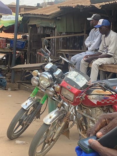 In Egbedi, Nigeria, a daylight scene depicts a street or market environment. Two motorcycles are parked prominently in the foreground on a dirt ground surface. The motorcycle on the left features chrome triple headlights, a chrome front fender, and green covers on its front shock absorbers. The red motorcycle on the right has a square headlight, a small windshield, and "DAYLONG" branding with a red, white, and blue checkered pattern on its fuel tank.
In the mid-ground, two men are seated on a wooden bench, facing right. The man on the left wears a light grey top and trousers with a black and white baseball cap; his arms are crossed. The man on the right wears a light-colored, possibly patterned, tunic and trousers with a blue and white baseball cap; he holds a small, light-colored object in his hands.
Behind the men and motorcycles, a rustic structure composed of wooden planks and corrugated metal sheets, with a rusty corrugated metal roof, functions as a stall or shop. Wooden tables are visible in front of this structure. To the far left, a green and white umbrella branded "Glo Cafe" partially covers a blue plastic crate and other items, suggesting another commercial point. In the immediate foreground, the dark-skinned hands of a person are visible, operating a black and green feature phone resting on a blue cloth.