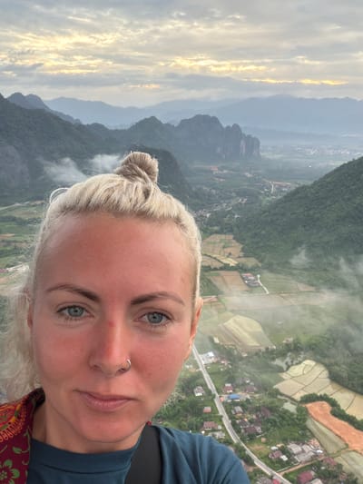 A selfie is taken from a high vantage point overlooking a valley in Hong Ngern Village, Laos. The foreground is dominated by the face of a blonde woman with blue eyes and a nose ring. Her hair is pulled up in a bun. In the background, a lush green valley is visible, with mist clinging to the mountains and a network of rice paddies and scattered buildings. The sky above is cloudy with hints of sunrise.
