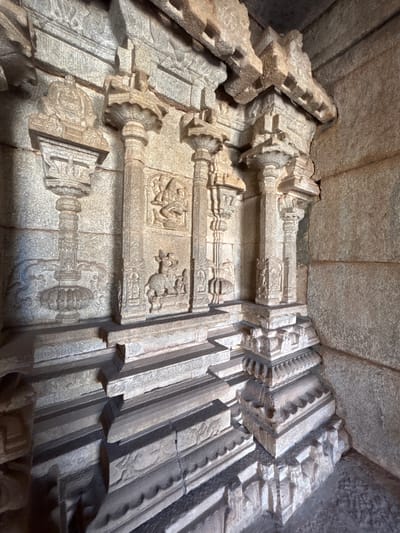 This image depicts the interior stonework of the Hazara Rama Temple, located in Kamalapura, India. The scene is an upward-angled view of sculpted walls and ceiling, showcasing detailed carvings in a light grey stone. The walls feature horizontal beams with decorative column-like structures. Carved depictions of figures and animals are integrated into the stonework. The architectural elements have a rough, aged texture. The ceiling is composed of plain stone blocks. Lighting within the scene casts shadows, highlighting the intricate details of the carvings.