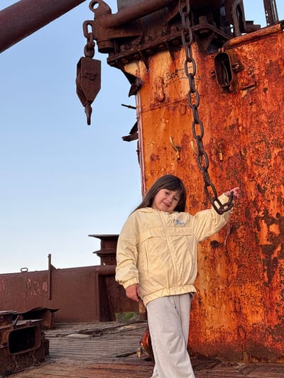 A young girl stands on the deck of a rusted ship in Teriberka, Russia. She is wearing a light yellow jacket and grey sweatpants, and she is smiling at the camera. The ship is heavily weathered, with rust covering its surfaces. A large chain hangs from a crane above the girl. The sky is clear and blue. The scene appears to be set in an outdoor environment, possibly a ship graveyard or a port.
