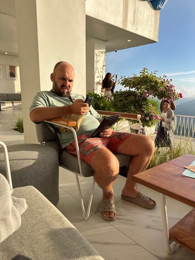 The image depicts a man seated in a grey chair on a patio. He has a bald head, a beard, and is wearing a light green shirt, patterned red shorts, and grey Crocs. He holds a black tablet in one hand and a phone in the other, glancing at the camera. He also wears a tan bag across his body. A brown wooden table is to his right.

In the background are two women, one smiling at the camera, standing near a bush with pink flowers. There is a white wall structure and a view of hills or mountains. The image appears to be taken in Kedisan, Indonesia, based on contextual clues.