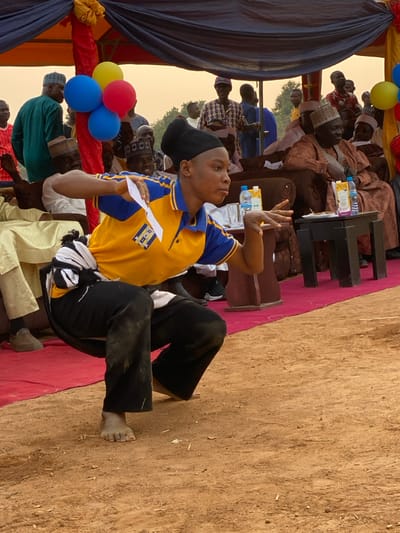 A person in a yellow and blue shirt, black pants, and bare feet sits on a red carpet. They are holding a white piece of paper and gesturing with their right hand. Behind them are seated people, some wearing traditional hats, and a black table with water bottles. Balloons decorate the scene. The background is a mix of draped fabric and dusty ground.