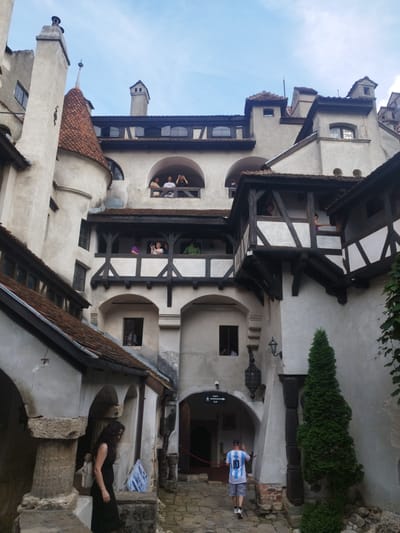 The image depicts the interior courtyard of Bran Castle, located in the city of Predeluț, Romania. The castle is characterized by its multi-story architecture, with cream-colored stucco walls, terracotta tiled roofs, and dark wooden beams supporting balconies and walkways. Several individuals are visible on balconies, appearing to observe the courtyard or take photographs. In the foreground, a woman in a black dress stands near stone pillars, and a person wearing a white jersey with the number "10" and the name "Messi" on the back walks away from the camera. A tall, conical evergreen tree stands to the right of the courtyard. The sky is visible in the upper portion of the image, with scattered clouds. The overall scene suggests a tourist visit to a historical landmark.