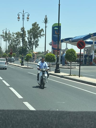 A photograph captures a street scene in Tassoultante, Morocco, featuring a man riding a light-colored scooter on a paved road. The man is wearing a light blue shirt. Behind him, there are trees, street lamps, and buildings, including a gas station. Cars and other vehicles are visible on the road and parked nearby. The sky is clear and blue. The road has white lane markings. A "no entry" road sign is visible in the background.