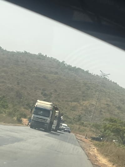 The image depicts a roadside scene in Assob, Nigeria, characterized by a busy two-lane highway flanked by a steep, sparsely vegetated hillside under an overcast or hazy sky. The perspective is from inside a vehicle, looking forward.
In the midground, a white semi-truck with a large cab and trailer is prominently positioned on the paved road, angled slightly to the left. Behind this truck, a queue of vehicles, including at least two additional trucks and several smaller passenger cars, extends down the road in a single lane.
To the left of the road, near the lead truck, several indistinct human figures are visible on the dirt shoulder. On the right side of the road, a dirt and rocky shoulder separates the pavement from the rising hillside. Further down the road on the right shoulder, a dark-colored vehicle, possibly a car, is stopped or parked. One or two additional human figures are faintly visible near this vehicle.
The background is dominated by a dry, brown hillside sparsely covered with scrub vegetation. A large, lattice-like electrical transmission tower stands prominently on the mid-upper slope of the hill on the right side of the frame, with faint power lines extending horizontally across the terrain.