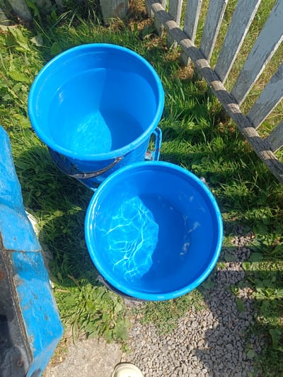 Two bright blue buckets filled with water sit on a grassy area next to a weathered wooden fence. The buckets are positioned close to each other, with the lower bucket slightly overlapping the upper one. Sunlight reflects off the surface of the water, creating shimmering patterns. Green grass and some weeds surround the buckets, with small gray gravel visible in the foreground. A portion of a blue painted object, possibly another container, is visible on the left edge of the frame. The scene appears to be outdoors, likely in a rural or backyard setting, given the grass, fence, and gravel. The lighting suggests it is daytime, possibly late morning or afternoon, with the sun casting shadows. There is no visible text in the image. The overall impression is one of a simple, everyday scene, perhaps related to watering plants or some outdoor activity.