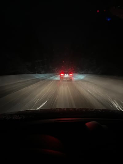 The image captures a nighttime scene on a snow-covered road in Arbuzovo, Russia, viewed from the perspective of a driver inside a vehicle. Heavy snowfall is actively illuminated by vehicle lights. A light-colored sedan is positioned ahead in the same lane, its red taillights illuminated. Headlights from the camera's vehicle cast prominent beams forward, intensely illuminating the falling snow particles which appear as bright streaks, and reflecting off the snow-covered road surface. The road itself is uniformly covered in snow, with faint disturbances visible where vehicles have passed. On both sides of the road, dark, indistinct masses suggest dense, snow-covered trees or vegetation under a completely dark sky. The lower portion of the image reveals the top of a vehicle dashboard and a human hand, possibly a right hand, resting on the dashboard area on the lower right, faintly illuminated with a reddish glow from inside the vehicle. Visibility is significantly impaired by the darkness and heavy snowfall.