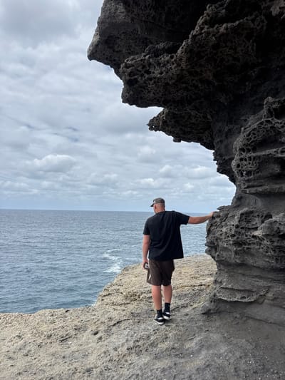 A man in a dark shirt, khaki shorts, and a baseball cap stands on a rocky ledge overlooking the ocean. He holds onto a jagged rock formation overhead. The sky, partly cloudy, is visible between the rock and the ocean. The rock formation has a dark, porous texture. The man appears to be posing for a photo.