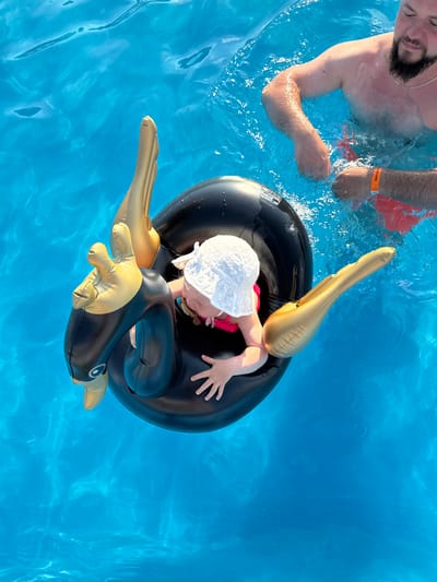 The image shows an overhead view of a person and a child in a bright blue swimming pool. The child is sitting in a black inflatable swan float with gold accents. The child is wearing a white hat and a pink swimsuit. To the right of the child, an adult male is partially visible in the water, wearing red swim trunks and an orange wristband. The water is clear and shows ripples. The setting is a swimming pool, possibly at a resort or private residence. The location is not explicitly shown, but the question specifies the city of Колокша, Russia.