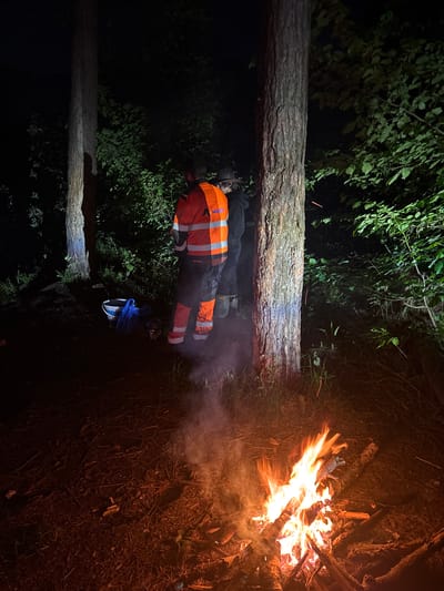 A man wearing an orange work vest and dark pants lies prone in the forest next to a fallen tree. His feet point towards a burning campfire. Nearby are some blue articles of clothing. The scene is dimly lit. The location is purportedly in Krasnyi Jar, Russia.