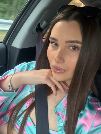 The image shows a young woman sitting in a car, with her head and shoulders visible. She has long, dark brown hair and brown eyes, and is wearing a colorful shirt with a pink and teal pattern. She is wearing a silver bracelet on her left wrist and sunglasses are perched on top of her head. Her right hand is resting on her chin. The interior of the car is visible, including the seatbelt and a portion of the car door. The exterior of the car appears to be a rural setting, with greenery visible through the window. The image was taken in Draganovtsi, Bulgaria.