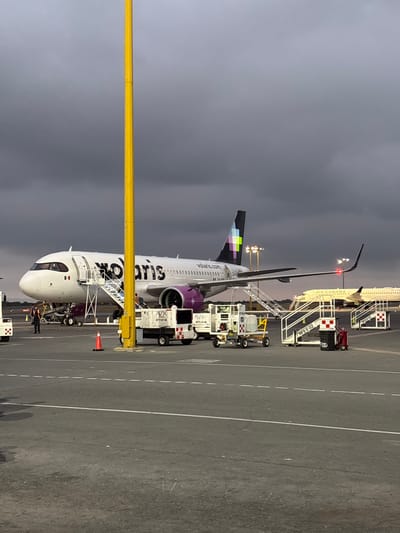 The image depicts a Volaris Airbus A320 aircraft parked on the tarmac at an airport, possibly in Apodaca, Mexico. The plane is white with purple accents and the word "volaris" prominently displayed. Passengers are disembarking via a set of stairs leading to the aircraft's front door. Ground crew members are visible near the plane. Ground support equipment, including a baggage loading vehicle and an air conditioning unit, are connected to the aircraft. In the background, another aircraft with stairs attached can be seen, labelled "Air Canada". The sky is overcast, suggesting a cloudy day or dusk. An orange traffic cone can be seen close to the airplane.