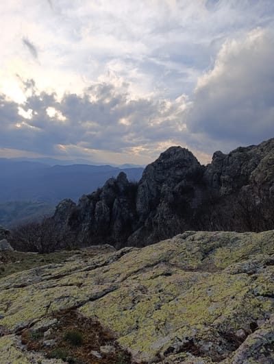 The image depicts a mountainous landscape in Ichera, Bulgaria. In the foreground, lichen-covered rocks dominate the view. Behind these rocks, a series of rugged, dark-colored rock formations are visible, punctuated by sparse, leafless trees. In the background, a series of layered, hazy blue mountains fade into the distance. The sky is filled with a mix of bright and dark clouds, with sunlight breaking through in certain areas. The overall light suggests either early morning or late afternoon. No people are visible.