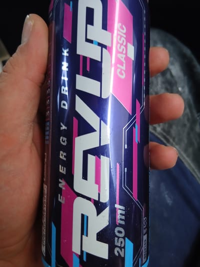 A hand holds a blue and pink energy drink can. The can has the brand name "REVOLVER" in large white letters, and "CLASSIC" and "ENERGY DRINK" are also visible in smaller text. The can also displays "250 ml". The background is out of focus, showing what appears to be dark fabric. There is no information in the image that indicates a location in Švenčionėliai, Lithuania.