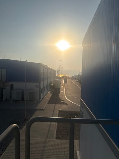 An eye-level, wide shot captures a scene bathed in the warm glow of the setting sun. The sun is positioned high in the sky, creating a lens flare that partially obscures the distant horizon. In the foreground, a grey metal railing runs across the bottom of the frame. To the left, a long, two-story modular building with blue upper walls and white lower walls stands parallel to a paved walkway. The number "02" is visible on the lower level of this building. To the right, the side of another large, blue modular building dominates the frame, its ribbed exterior reflecting the sunlight. The walkway extends into the distance, where it is lined with more modular structures and streetlights. A truck is visible in the distance on the walkway. The overall environment appears to be an industrial or temporary housing complex. The location is identified as Gülnar, Turkey.