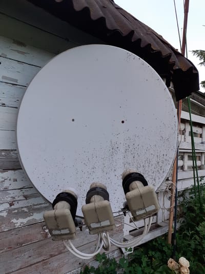 A satellite dish with three LNBs is mounted on a weathered wooden wall of a building, possibly a shed or outbuilding, in Deremezna, Ukraine. The dish is white and appears to be covered in light dust or dirt. The LNBs are beige and have black collars. White coaxial cables connect to the LNBs and trail downwards. The building's wall is made of horizontal wooden planks, painted a light blue or green, with peeling paint revealing older layers and the wood itself. A rusty corrugated metal roof is visible above the dish. The background shows more of the building with white wooden structures, green vegetation, and a pale sky, suggesting it is daytime. There are no people or animals visible in the image.