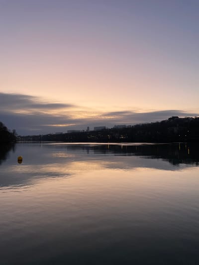 The image displays a serene twilight or dawn scene over a calm body of water, likely a river or lake, near Caluire-et-Cuire, France. The scene is bisected horizontally by a distant shoreline, with the sky above and reflections on the water below.
The sky, occupying the upper portion, transitions from a soft, pale purple on the left to a warmer, gentle yellow-orange hue on the right, indicating the soft light of sunrise or sunset. Wispy clouds are present, mirroring these subtle colors.
Along the distant horizon, a silhouette of a landmass is visible, densely covered with dark trees and structures. Numerous small, artificial lights dot this shoreline, glowing brightly against the receding daylight or approaching night.
The surface of the water is remarkably still, acting as a near-perfect mirror. It clearly reflects the nuanced colors of the sky and the twinkling artificial lights from the distant buildings. Subtle ripples on the water's surface add texture to these reflections.
In the lower right section of the water, a distinct yellow buoy with a black horizontal band is present. Its reflection is also faintly visible beneath it. The overall atmosphere is tranquil and peaceful, showcasing the beauty of the natural environment at the transition point of day. No visible text is present in the image.