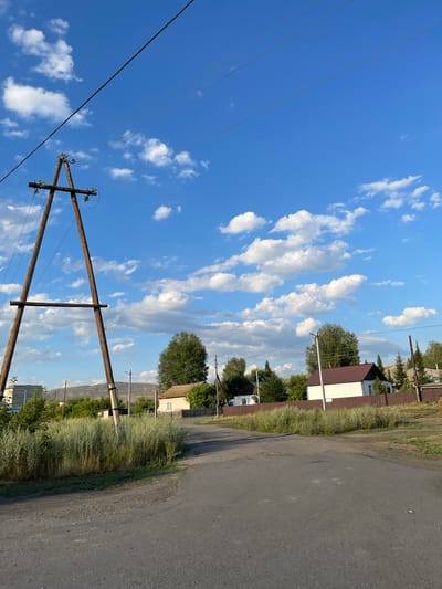 The image shows a rural street scene in Predgornoe, Kazakhstan, under a bright blue sky with scattered white clouds. A wooden power line pole, constructed from two angled posts joined by a horizontal beam, stands prominently on the left side of the road. Power lines stretch across the upper portion of the frame.
The road itself is unpaved and appears to be a dirt or gravel track, with some patches of asphalt visible. To the right of the road, a row of houses with dark brown roofs and white walls are visible, separated by a brown wooden fence. Green trees and tall, dry grass are abundant throughout the scene. In the background, hills or mountains can be seen in the distance. The overall impression is one of a quiet, rural settlement.