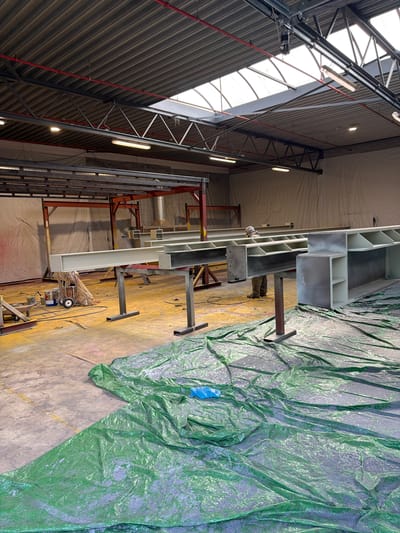 The image depicts an indoor industrial setting, likely a workshop or manufacturing facility in Mol, Belgium. The scene is dominated by large, light-colored steel beams, some of which appear to be newly fabricated or painted, resting on metal stands. A person, dressed in work clothes and a hooded protective suit, is visible working on one of the beams. The floor is a mix of concrete and yellow-stained surfaces, with a large green tarp spread out in the foreground, covered in dust or debris. Various tools and equipment, including what appears to be a paint sprayer and buckets, are scattered around. The overhead structure of the building, with its metal beams, corrugated roof, and skylights, is also visible, suggesting an industrial workspace designed for large-scale projects. The overall impression is one of ongoing construction or fabrication work.