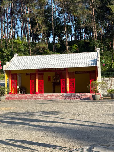 The image shows a long, low building with a white tiled roof and red pillars. The building is located in Lao Cai Ward, Vietnam. The road is to the side of the building, and trees are in the background. The building has a sign in Vietnamese that reads "KHÔNG GIAN VĂN HOA TRÂN TRIEU".
