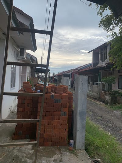 The image depicts a street scene in Blangbladeh, Indonesia. A large stack of red bricks sits near a concrete wall along the edge of the street. A metal ladder-like structure partially obscures the view of the bricks and adjacent buildings. Beyond the bricks, a row of houses and trees line the street. Overhead, the sky is visible with some cloud cover. The environment suggests a residential area undergoing construction or renovation.