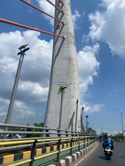 The image shows a concrete pylon of a bridge, with red cables attached to it. The pylon has a sign that reads "JEMBATAN SEI ALALAK". There are streetlights attached to metal poles on the side of the road. The road is asphalt and has a yellow and black striped barrier. Several motorcycles are visible on the road, with some people wearing helmets and backpacks. The sky is blue with white clouds. The location is identified as Berangas Timur, Indonesia.