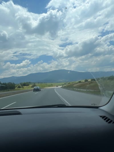 The image shows a view from inside a car driving on a highway. The highway has multiple lanes and is bordered by a grassy median and a guardrail. On the left side of the highway, there are trees and a green field. In the distance, there is a mountain range with a hazy blue hue, suggesting it is a considerable distance away. The sky is partly cloudy with fluffy white clouds against a blue sky. There are two cars visible on the highway, one in the left lane and another further ahead in the right lane. The foreground of the image shows the dashboard of the car, with a portion of the windshield and the windshield wipers visible. The image is taken from the perspective of the driver or a passenger. The overall impression is a scenic drive on a highway in a mountainous region on a partly cloudy day. The location is Golemo Buchino, Bulgaria.