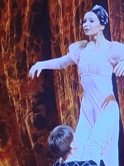 In the city of Bolshoye Afanasovo, Russia, a ballet performance is captured in a photograph. A female ballet dancer, dressed in a light pink, floor-length gown with puffed sleeves and intricate bodice detailing, is center stage. She wears white gloves and her hair is styled in an elaborate updo with a decorative hairpiece. Her facial expression is serious, and her body is posed in a graceful dance stance. She is being supported by another performer, partially visible at the bottom of the frame, whose back is turned to the camera. The backdrop consists of heavy, textured curtains in shades of orange and brown, creating a warm, dramatic atmosphere. The lighting appears to be theatrical, highlighting the performers and the stage setting.
