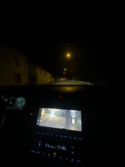It's nighttime, inside a car with dashboard lights illuminating the console. A rearview camera displays a white car behind. The outside view is visible through the windshield, with streetlights lining the road. Dashboard readings show a temperature of 20.5 degrees Celsius and a speed of 65 km/h. The console has buttons for volume, navigation, radio, media, and setup, alongside air conditioning controls and synchronization options.