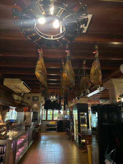 The image shows the interior of a shop selling cured meats, likely a charcuterie or delicatessen. Several large, cured ham legs are hanging from the wooden ceiling. The shop features a display counter with refrigerated sections showcasing various meat products. In the background, there's a person in a white shirt and black apron standing by a window. The environment appears to be a commercial establishment with wooden beams, tiled floors, and multiple display cases and refrigeration units. One of the refrigeration units has the logo "RAMON" on it. The image does not provide information about the city of Gènova, Spain.