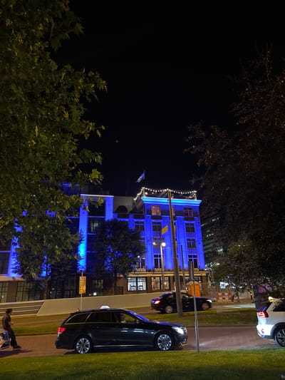 This media file captures a nighttime scene in Arnhem, Netherlands. The primary subject is a modern building illuminated with vibrant blue lights, creating a striking contrast against the dark sky. The building appears to be multi-storied, with visible windows reflecting the blue light. Decorative string lights are draped across the facade of the building.
In the foreground, a lush green lawn runs alongside a paved walkway. Several cars are parked or driving on the adjacent road. A dark-colored sedan is prominently featured, reflecting the blue illumination. Further down the road, a white car is also visible.
Trees with dark foliage are scattered throughout the scene, some partially obscuring the building. The time of day is clearly night, and the weather appears calm. The overall ambiance is urban and contemporary, with a festive or atmospheric feel due to the lighting.
A person is seen in the lower right corner of the frame, sitting on a motorcycle and appearing to be holding something, possibly food or a drink. They are dressed in dark clothing. The scene suggests a moment of quiet observation or waiting amidst the urban environment. There is a yellow sign with text visible on a pole near the cars.
