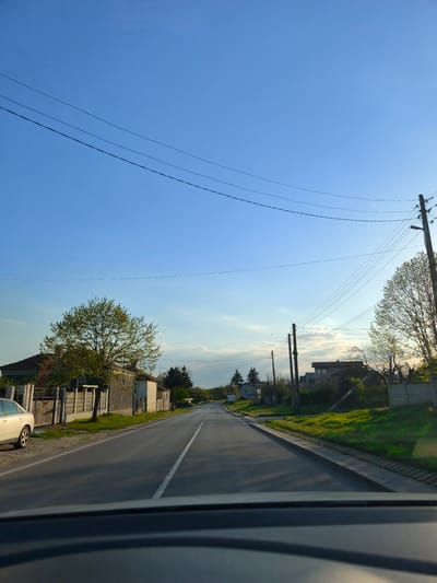 Captured from within a vehicle, the photograph depicts a residential street in Zdravets, Bulgaria. The asphalt road is characterized by a single white lane marker bisecting its width. On the left, a parked silver-colored car faces forward, next to fenced houses with trees in the yard. Electrical poles and wires traverse the sky above. The sky is blue. The scene is captured in daylight, but the lighting is beginning to indicate the oncoming evening. The image is taken from the driver's position.