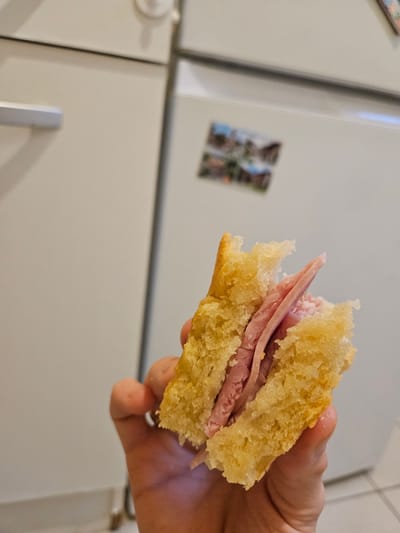 A person holds a partially eaten ham sandwich in their right hand. The sandwich consists of two pieces of yellow bread, each with a slightly irregular shape, containing multiple slices of ham. The background features a white refrigerator with a vertical silver handle on its lower section, and a photograph magnet is visible on the upper section. The location is Villeneuve-Saint-Georges, France is not verifiable from the image.