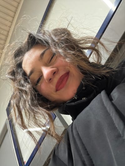 A woman with fair skin and curly brown hair tilts her head downwards, her eyes mostly closed. She wears dark lipstick. A black jacket with a high collar is visible. The background features a slightly blurred, light-colored surface with blue and white details. The photo's angle suggests it was taken from a high perspective, looking down at the subject.